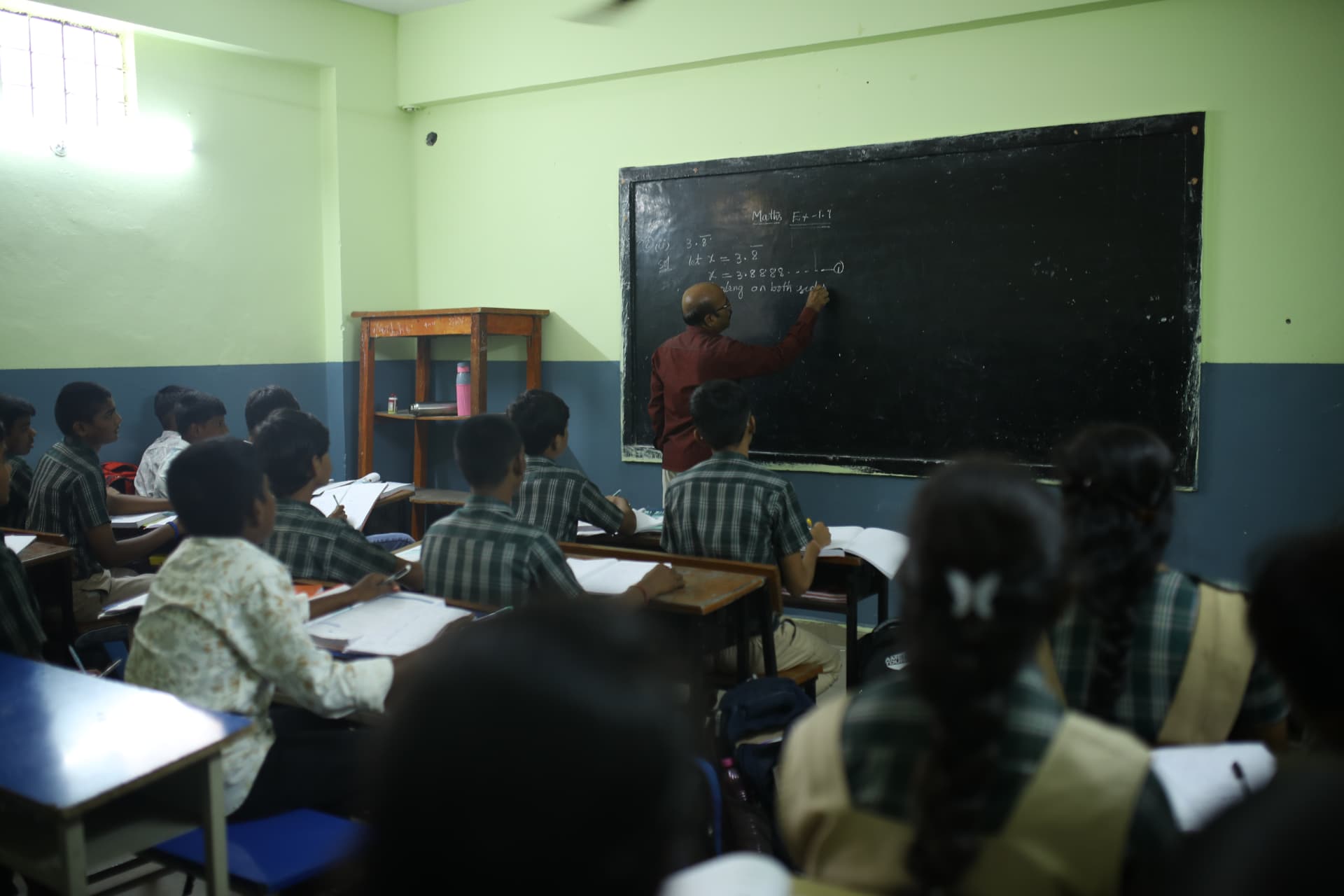 A teacher writing a mathematics lesson on the blackboard while students follow from their desks