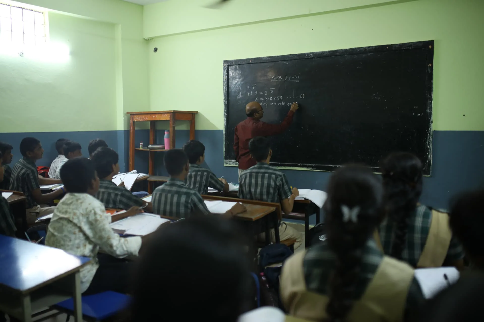 A teacher writing a mathematics lesson on the blackboard while students follow from their desks